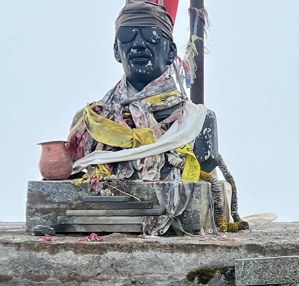 Bust of Sudip Gautam at Badal Danda on the Mardi Himal trail, draped in khata scarves and a marigold garland, with a copper offering vessel beside it in fog