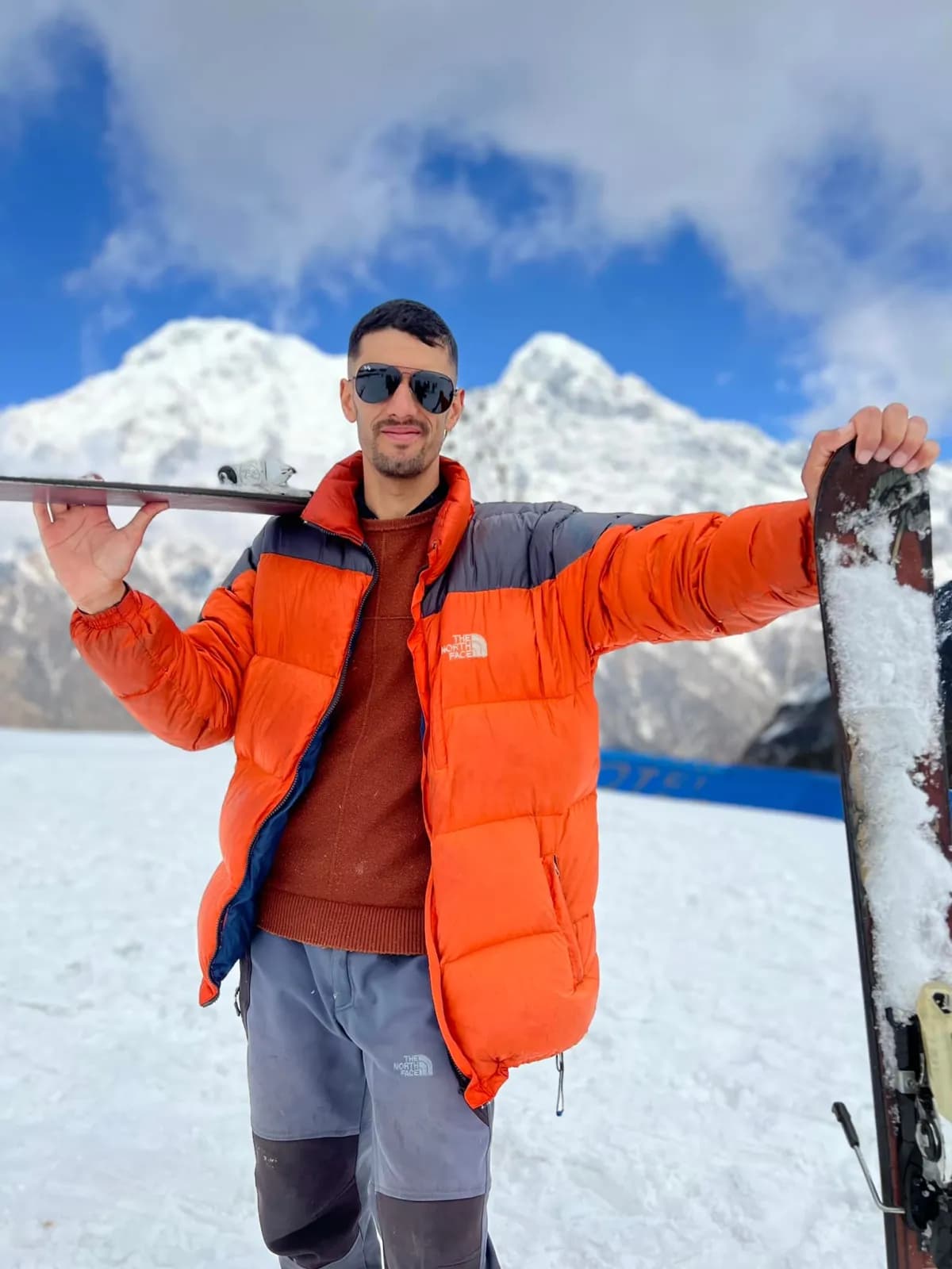 Sudip Gautam in an orange down jacket and aviator sunglasses, holding a pair of skis in the snow below a white Himalayan peak