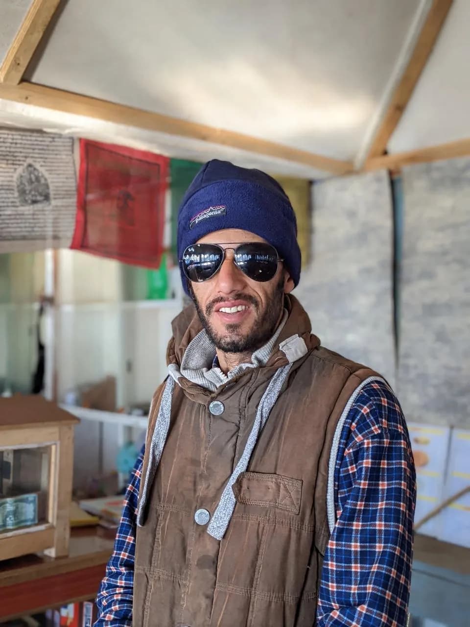 Sudip Gautam in a blue beanie, aviator sunglasses and a brown vest, smiling inside a teahouse with prayer flags on the wall